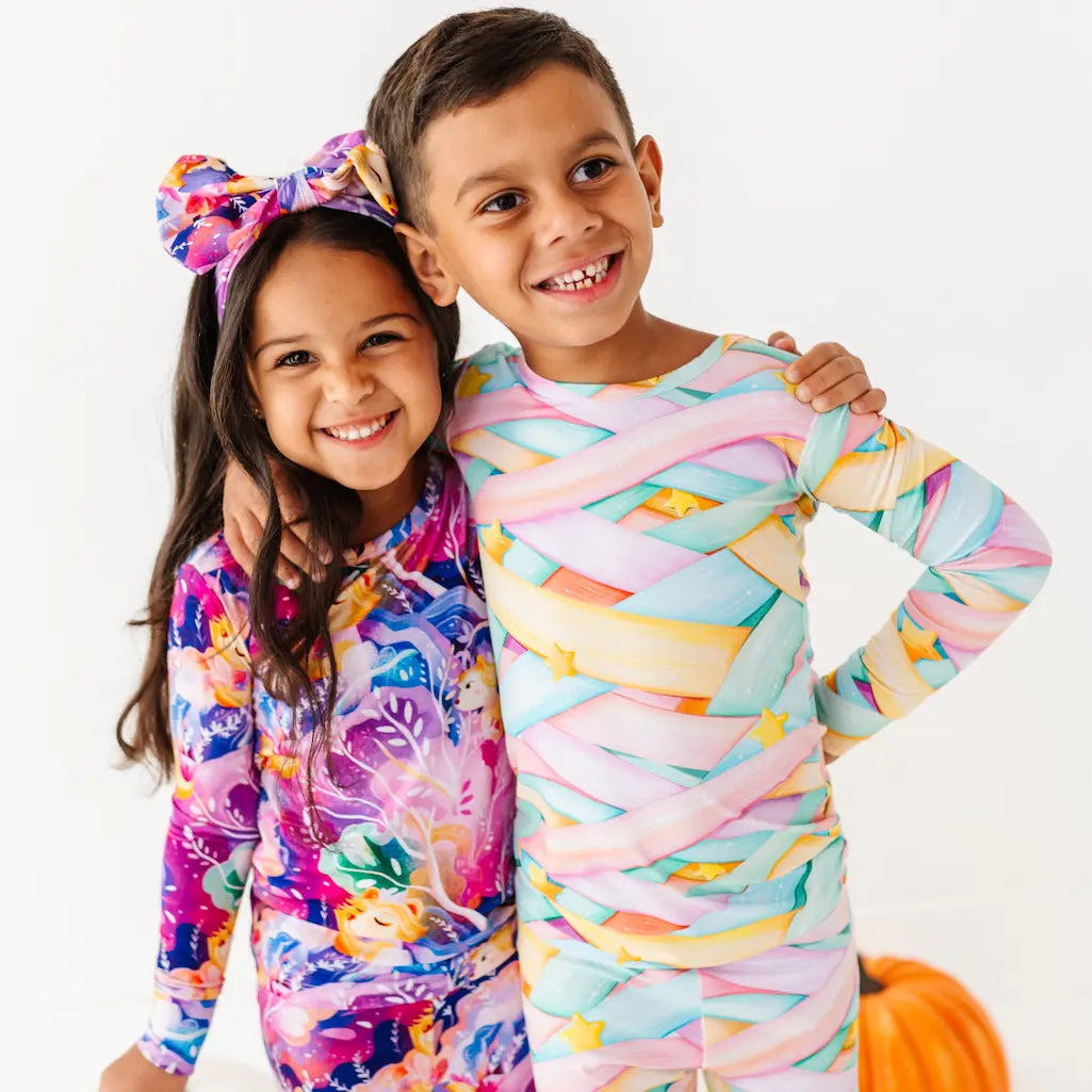Two children standing together and smiling, one wearing a purple and multicolored sea-themed pajama set with a large matching bow headband, and the other in pastel candy ribbon-patterned pajamas, with small pumpkins on a white background.