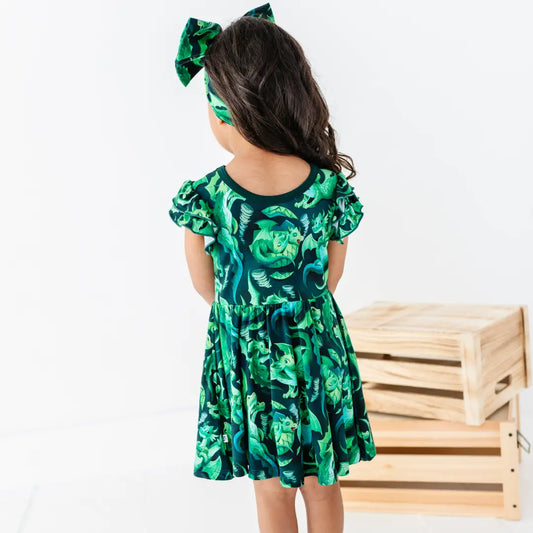 Child wearing a green dress with a leaf pattern and matching headband, standing against a white background.