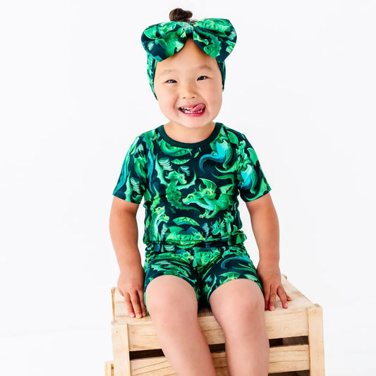Child wearing a green dinosaur print outfit sitting on a wooden block against a white background