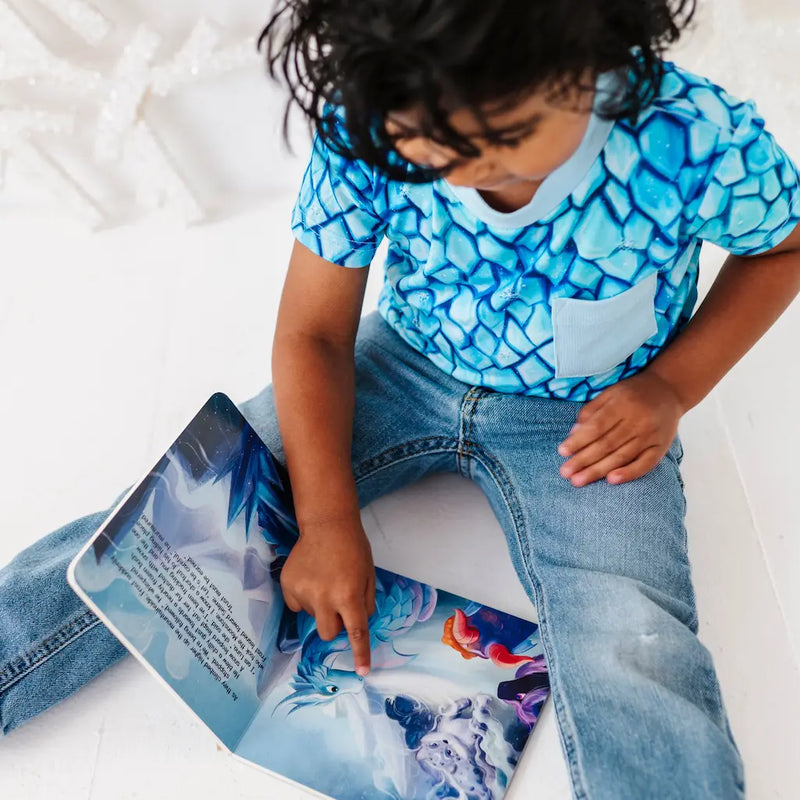 Child wearing a blue patterned shirt and jeans, sitting on a white surface with a colorful book.