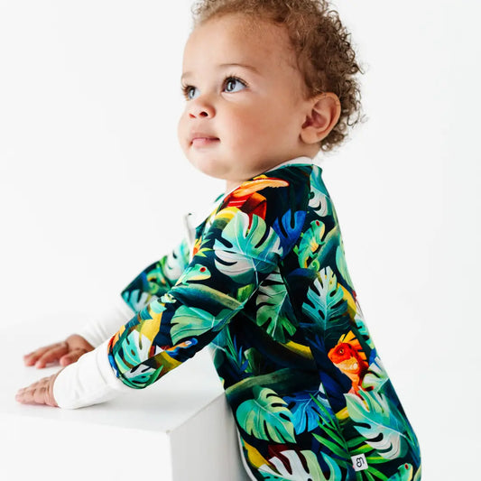 Curly-haired toddler leaning on a white block and looking up, wearing a green tropical jungle-print onesie with iguanas and leaves and white cuffs, on a white background