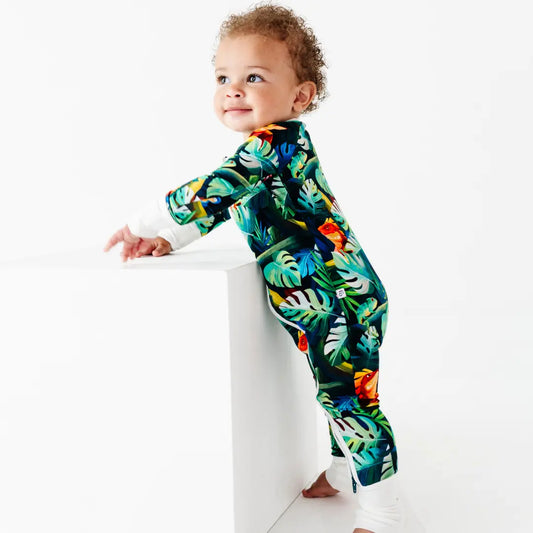 Curly-haired toddler leaning against a white block, wearing a green tropical jungle-print onesie with iguanas and leaves and white cuffs, on a white background