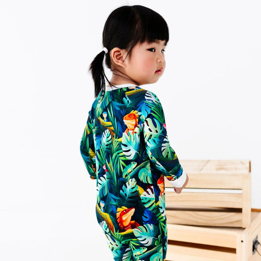 Young girl with a ponytail seen from behind looking over her shoulder, standing next to a wooden crate, wearing a green tropical jungle-print onesie with iguanas and leaves, on a white background