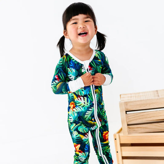 Smiling young girl with pigtails standing next to a wooden crate, wearing a green tropical jungle-print zip-up onesie with white trim, on a white background