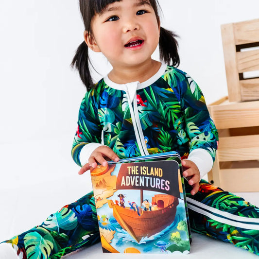 Young girl with pigtails sitting and holding a children's book titled The Island Adventures, wearing a green tropical jungle-print onesie, next to wooden crates