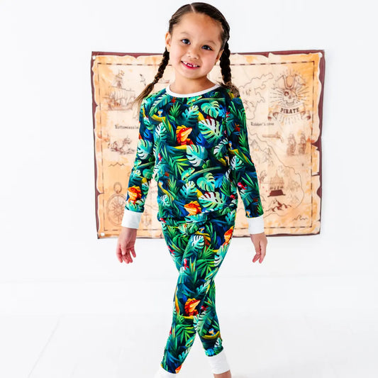 Smiling young girl with braided pigtails standing and wearing a green tropical jungle-print pajama set with parrots and leaves, with a treasure map decoration on the wall behind