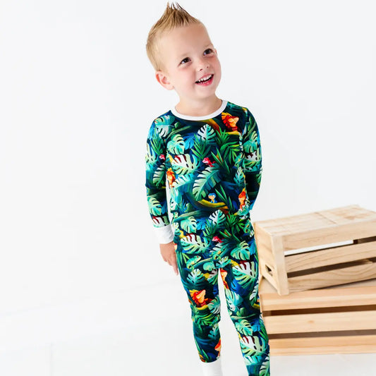 Smiling young boy with spiky blond hair standing next to wooden crates, wearing a green tropical jungle-print pajama set with iguanas and leaves and white cuffs and collar, on a white background