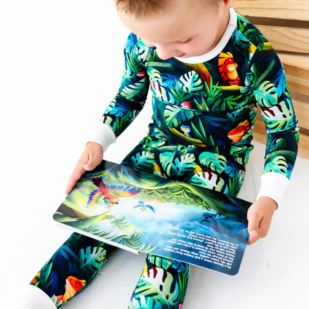 Young boy sitting and reading an illustrated children's book showing a parrot in a jungle scene, wearing a green tropical jungle-print pajama set with iguanas and leaves and white cuffs