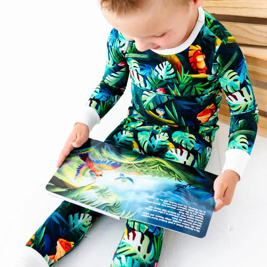 Young boy sitting and reading an illustrated children's book showing a parrot in a jungle scene, wearing a green tropical jungle-print pajama set with iguanas and leaves and white cuffs