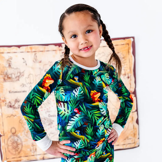 Smiling young girl with braided pigtails standing with hands on hips, wearing a green tropical jungle-print pajama top with iguanas and leaves, with a treasure map decoration on the wall behind