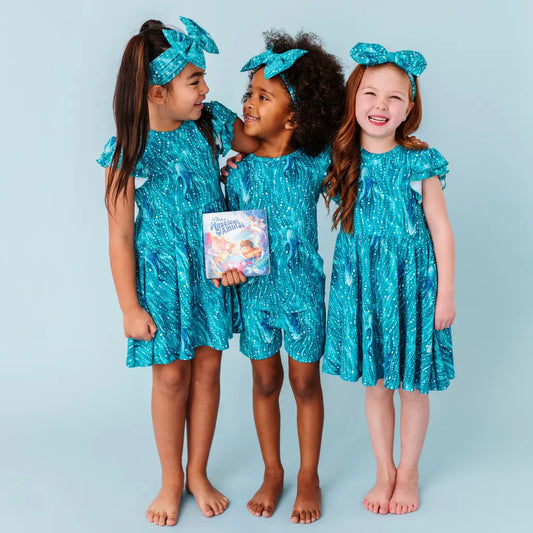 Three children wearing matching blue dresses with a pattern, standing against a light blue background.