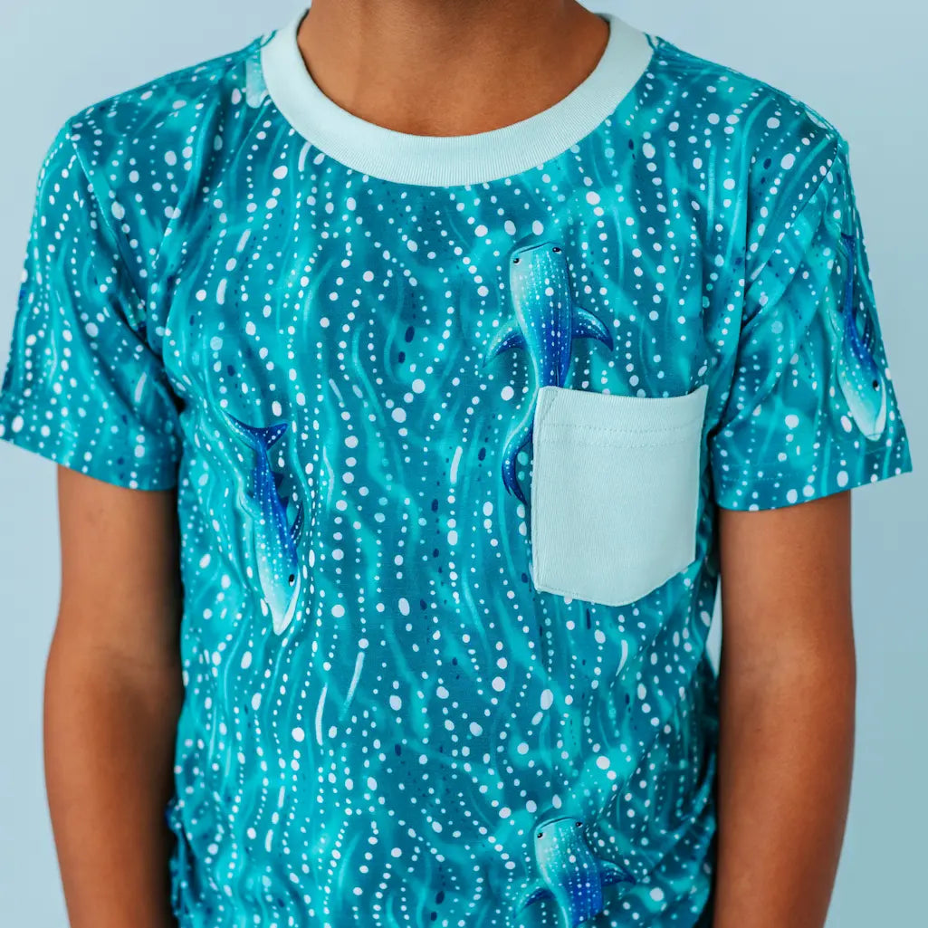 Child wearing a teal whale shark patterned t-shirt with a white pocket on a light blue background