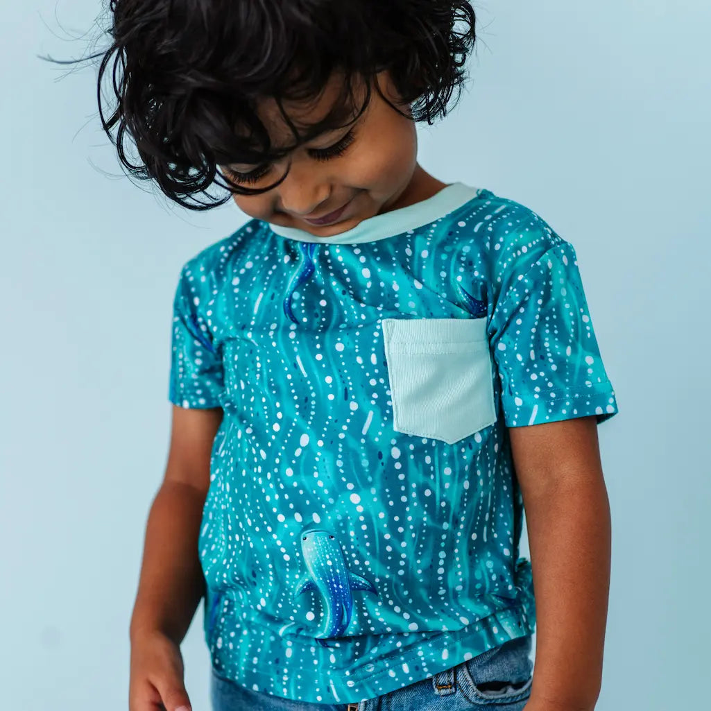 Child wearing a teal whale shark patterned t-shirt with a white pocket on a light blue background