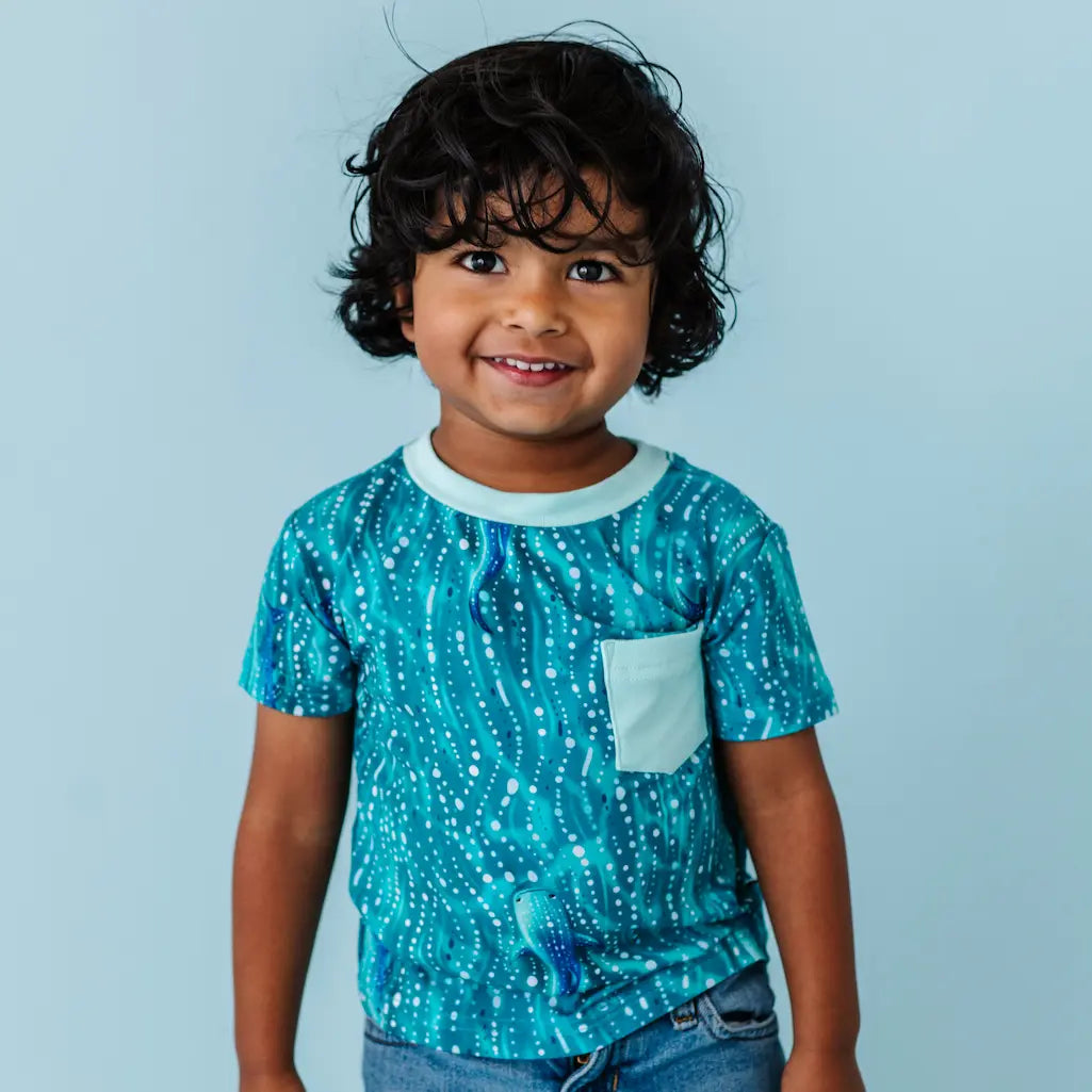 Smiling child wearing a teal whale shark patterned t-shirt with a white pocket on a light blue background