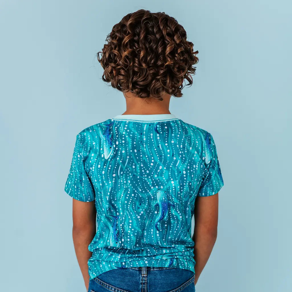 Back view of a child wearing a teal whale shark patterned t-shirt on a light blue background