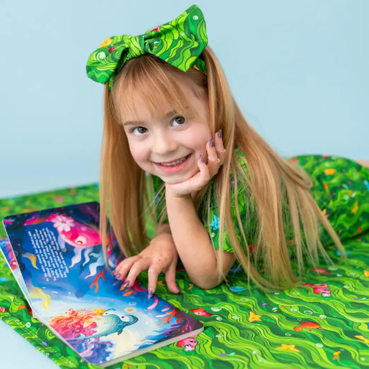 Child in a green dress with a matching headband, reading a book on a green surface.