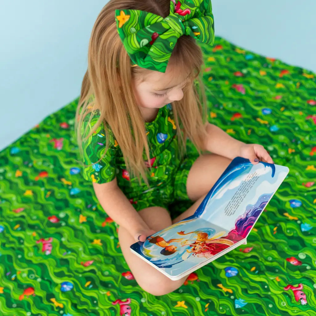 Child in a green outfit with a matching headband reading a book on a green mat with fish patterns.