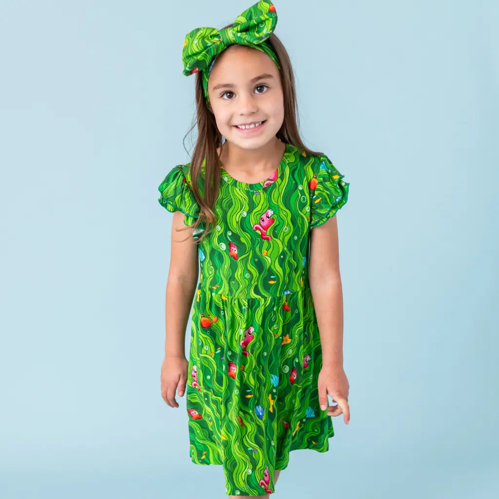 Young girl wearing a green dress with a pattern and matching headband against a light blue background