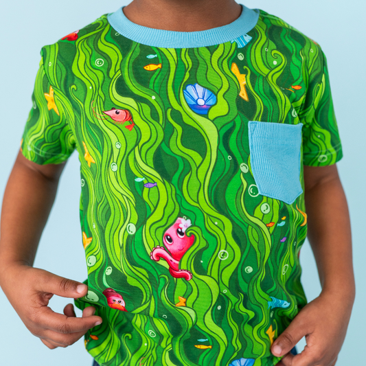 Child wearing a green underwater-themed t-shirt with fish and shell designs on a light blue background