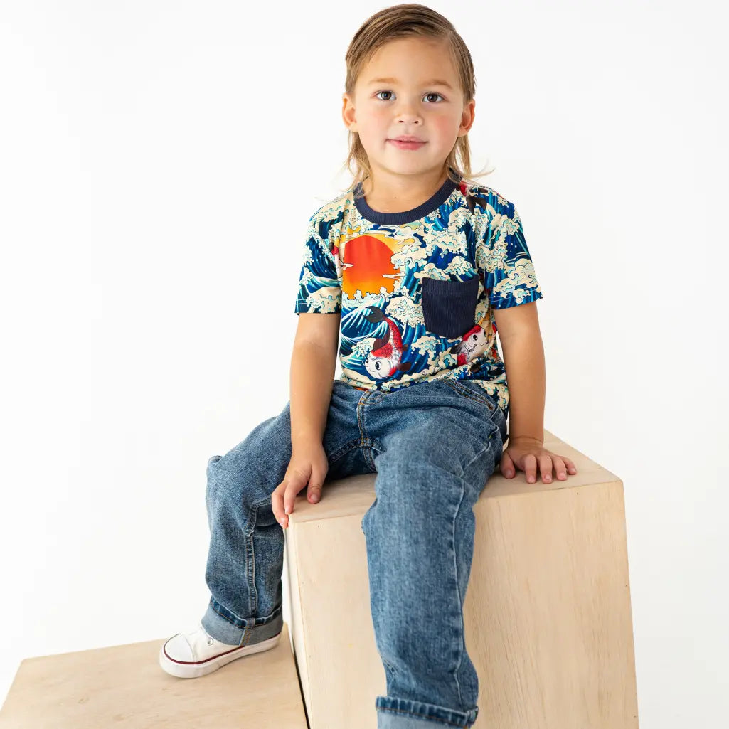 Child sitting on a block wearing a colorful t-shirt and jeans against a white background