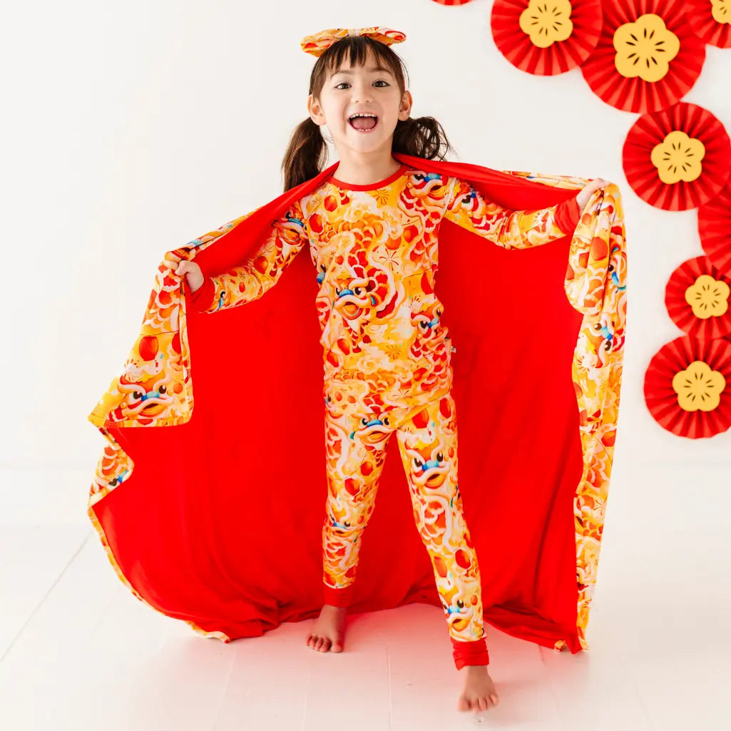 Child wearing a colorful outfit with a red cape, surrounded by decorative red and yellow elements on a white background.