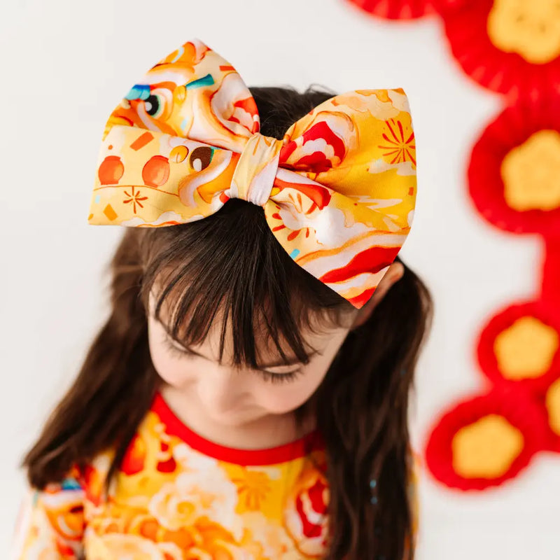 Child wearing a large colorful bow with a red and yellow flower in the background