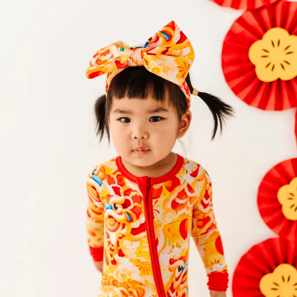 Child wearing a colorful outfit with a large bow, standing against a white background with red floral decorations.