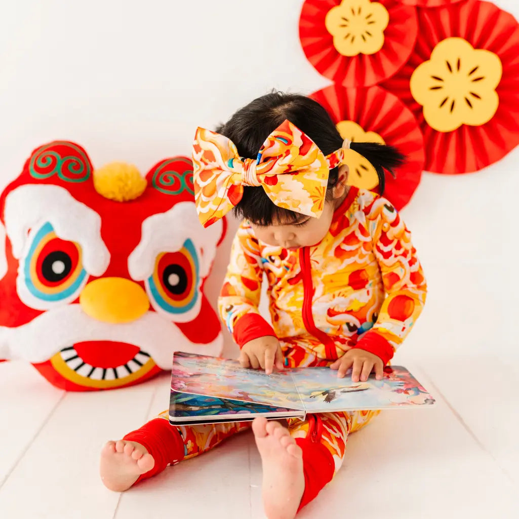 Child in a colorful outfit with a large bow reading a book, with festive decorations in the background.