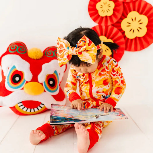 Child in a colorful outfit with a large bow reading a book, with festive decorations in the background.