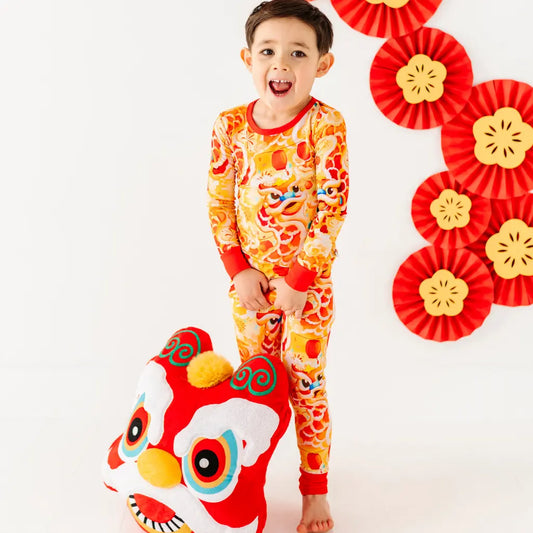 Child wearing a colorful pajama set with a dragon design, standing next to a red dragon plush toy on a white background.