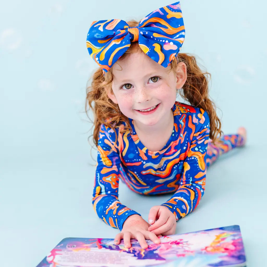 Child wearing a colorful tie-dye outfit with a large bow, lying on a light blue background.
