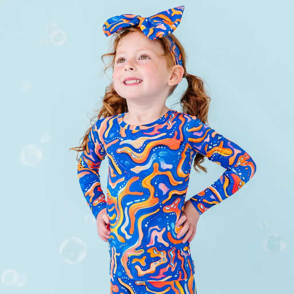 A child wearing a long sleeve top and matching pants with a vibrant blue and orange swirling pattern, resembling a mandarin fish. The child also wears a matching headband.