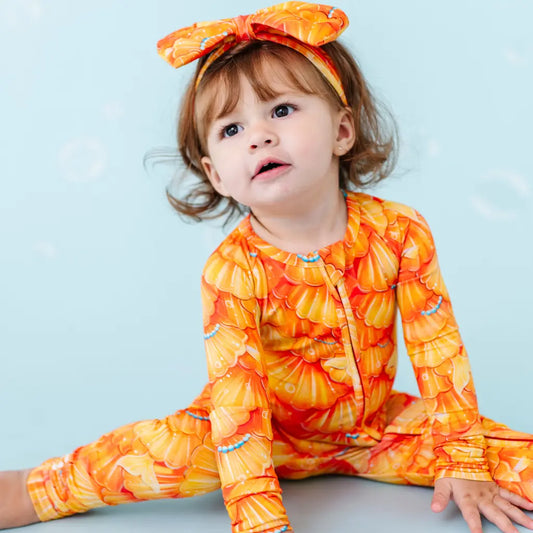 A baby wearing a one-piece zipper pajama with a mermaid pattern of golden seashell scales and turquoise pearls, matching bow on head.