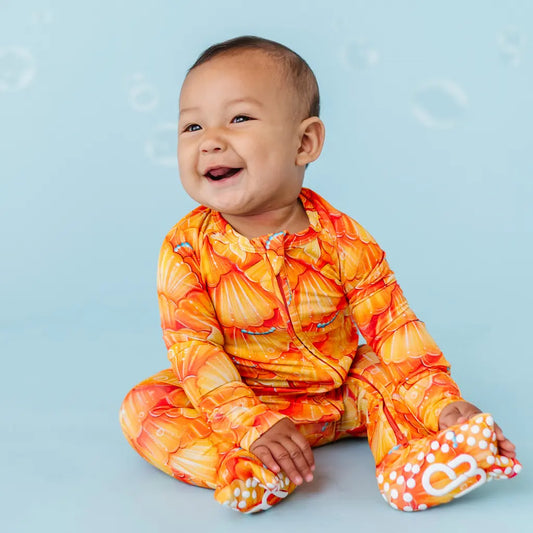 A baby sitting on a blue surface wearing a one-piece zipper pajama with a mermaid pattern of golden seashell scales and turquoise pearls.
