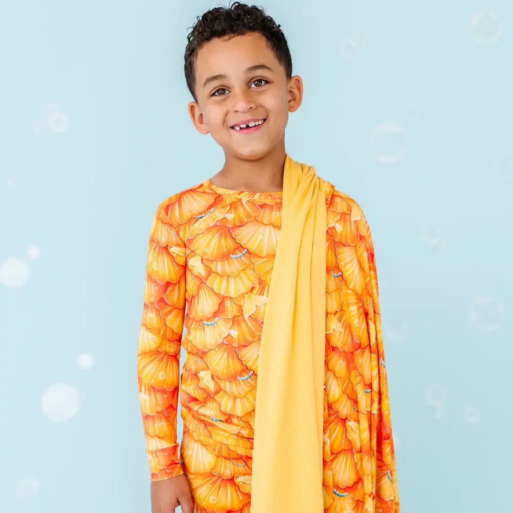 A child standing against a blue background with sparkles, wearing a orange blanket with a mermaid scale pattern and a yellow scarf.