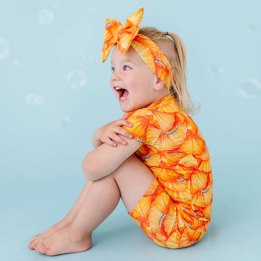 A child sitting on a blue background, wearing a short pajama set with a mermaid pattern in orange and white tones, and a matching headband.