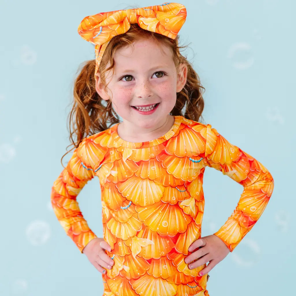 A child wearing a pajama set with a mermaid scale pattern in orange and yellow tones, complemented by a headband with a bow.