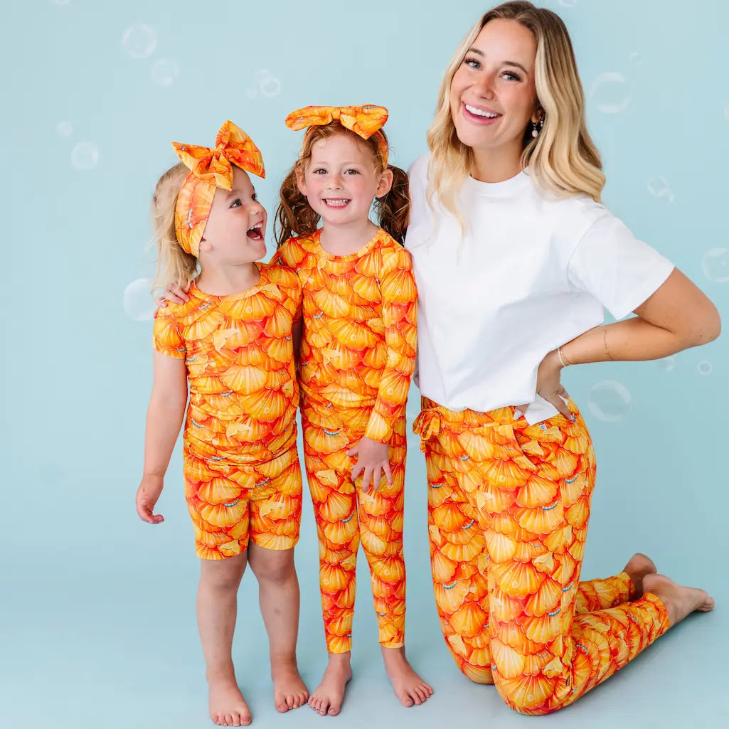 Woman and two children in matching orange and yellow pajama outfits on a light blue background.