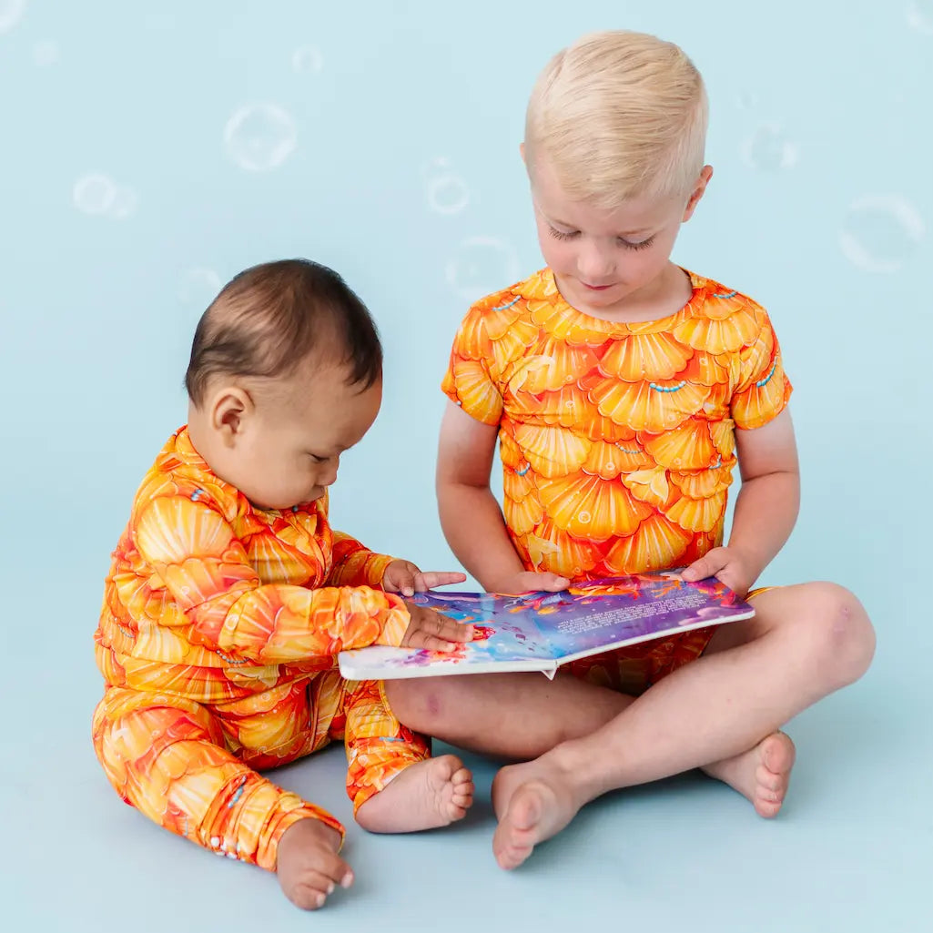 Two children in orange and yellow swimsuits sitting on a blue background reading a book.