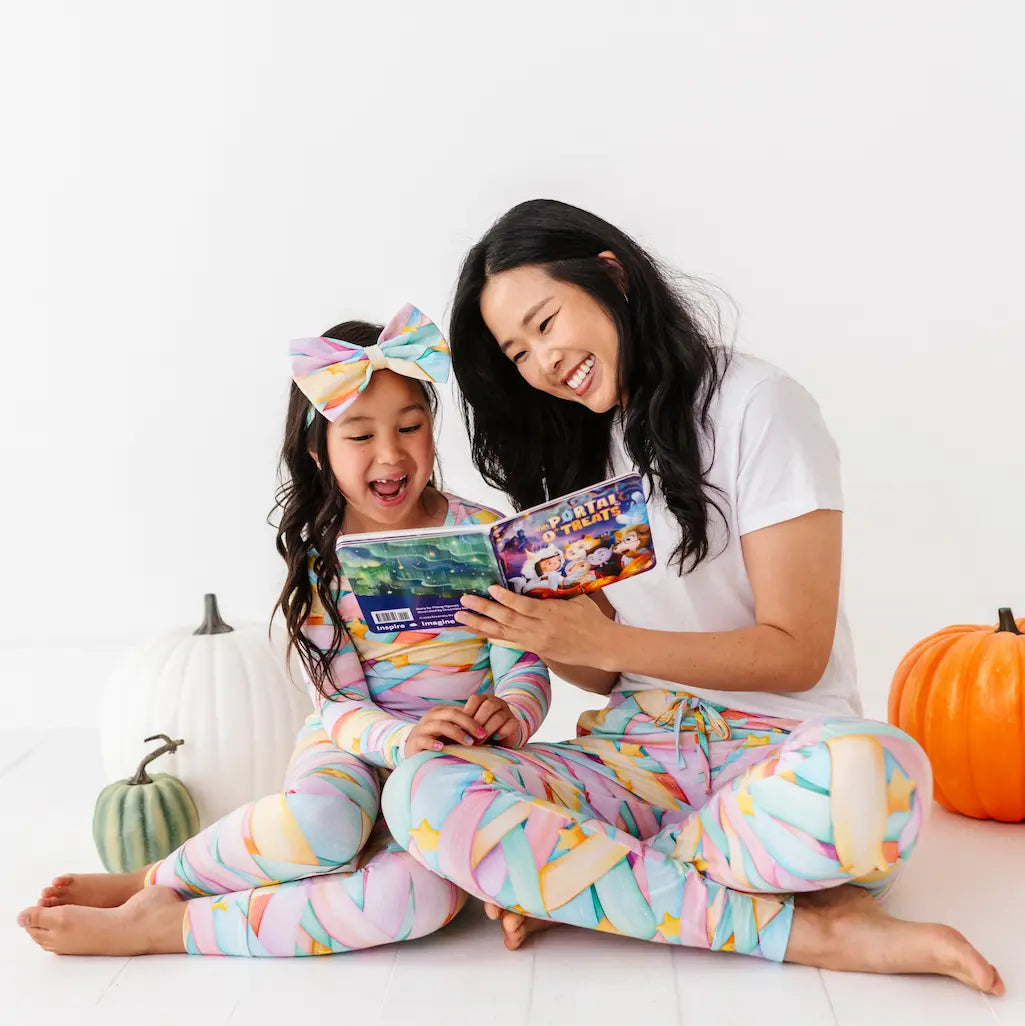 Mother and daughter wearing matching pastel candy ribbon-patterned pajamas, the child with a large matching bow headband, sitting barefoot on a white floor and reading an illustrated book together with pumpkins nearby against a white background.