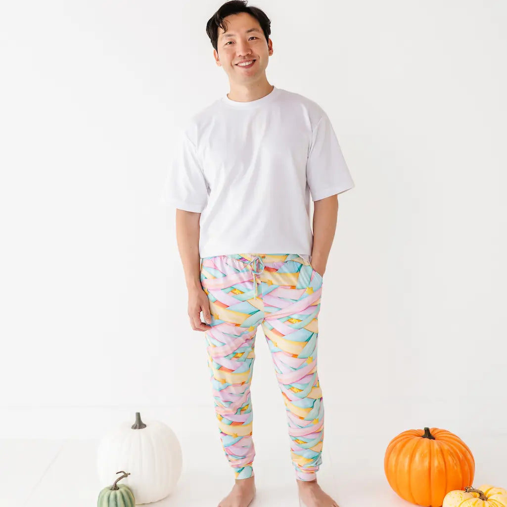 Smiling man in a white T-shirt wearing colorful pastel candy ribbon-patterned pajama pants with a drawstring and yellow stars, standing barefoot next to pumpkins on a white background.