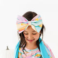 Child wearing a colorful oversized pastel headband bow with yellow stars and a matching pastel-patterned outfit, looking down against a white background.