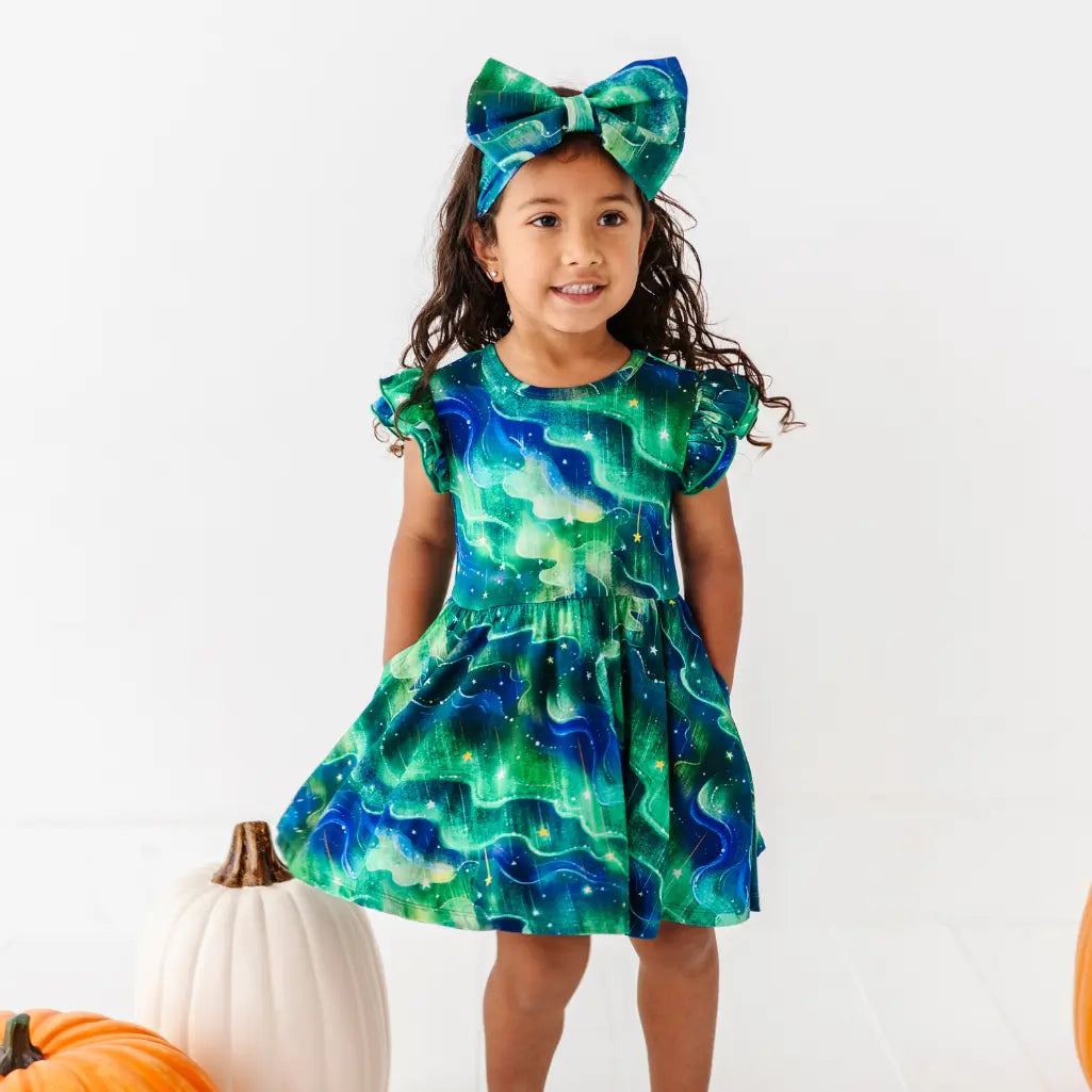 Young girl wearing a green and blue dress with a matching bow, standing against a white background.