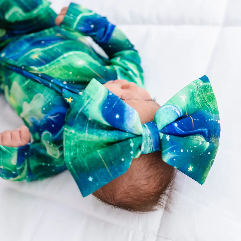 Baby wearing a green and blue outfit with a large bow, lying on a white blanket.