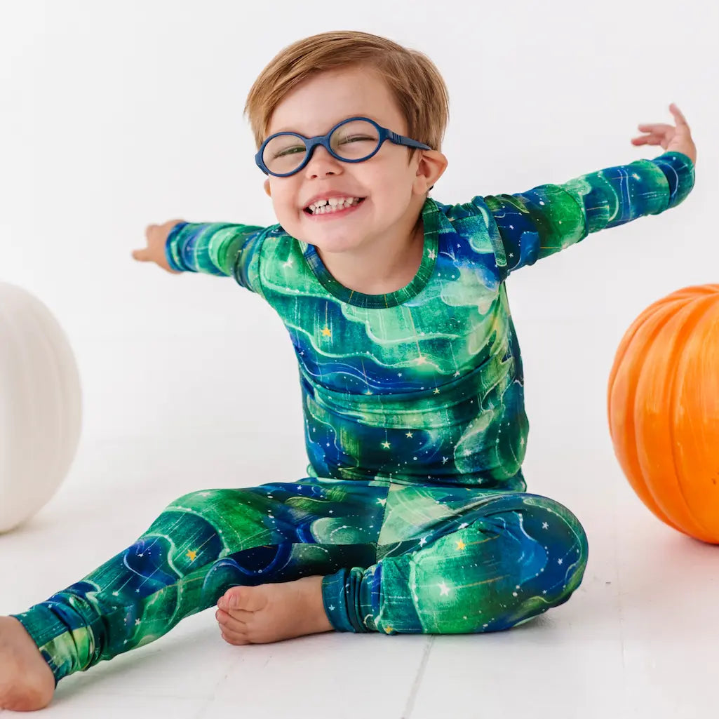 Child wearing a green and blue patterned outfit with glasses, sitting on a white floor with orange and white balls.