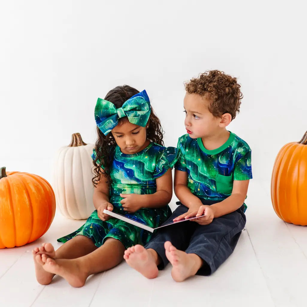 Two children in matching green and blue outfits with pumpkins on a white background