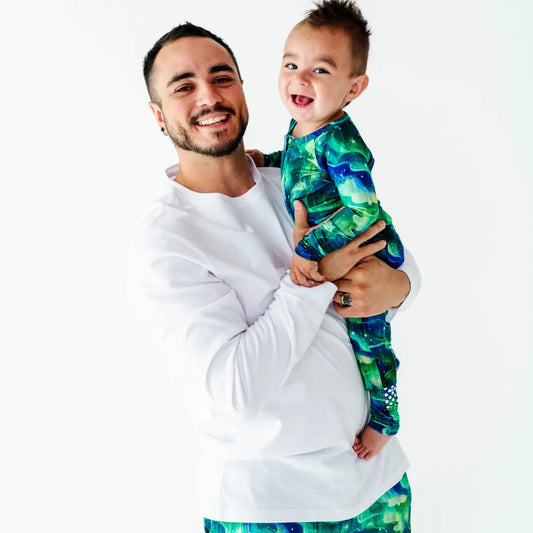 Smiling man in a white top holding a laughing baby who is wearing a green and blue galaxy-patterned pajama, on a white background
