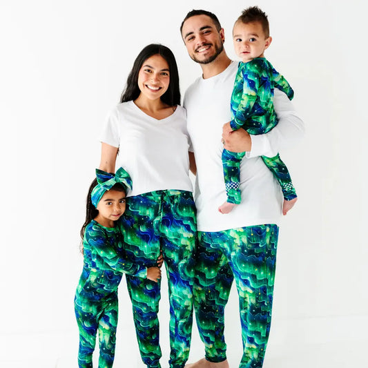 Smiling family of four standing together, the parents in white tops with green and blue galaxy-patterned bottoms and the two children in matching green and blue galaxy-patterned pajamas, on a white background