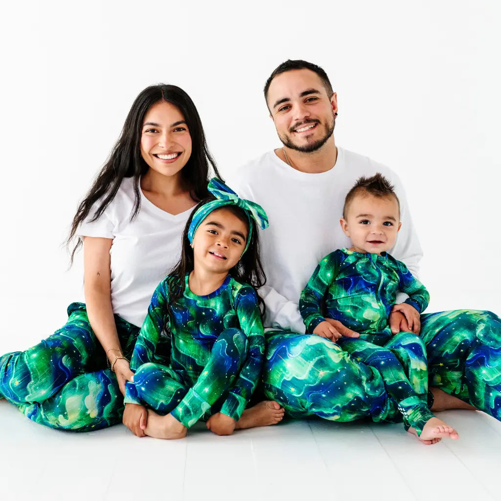 Smiling family of four sitting together on a white floor, the parents in white tops with green and blue galaxy-patterned bottoms and the two children in matching galaxy-patterned pajamas, on a white background