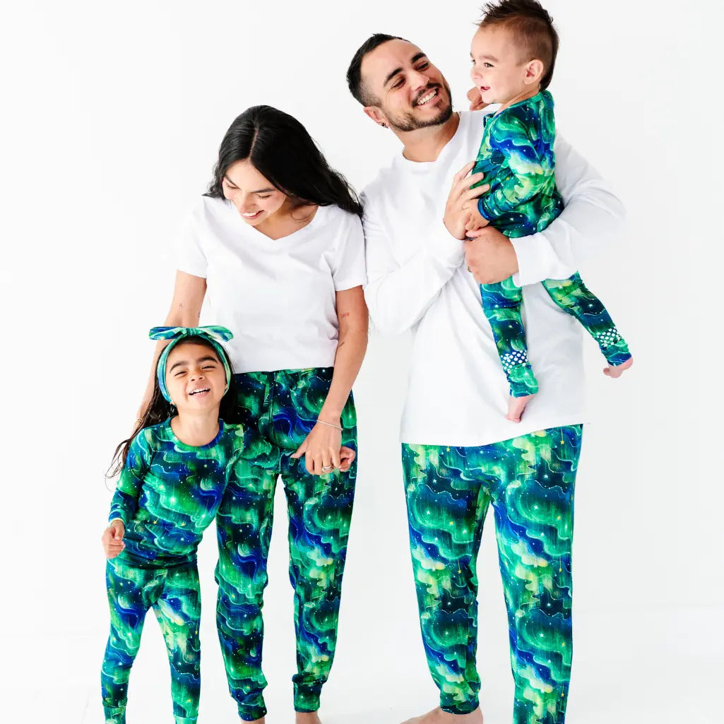 Happy family of four standing together and laughing, the parents in white tops with green and blue galaxy-patterned bottoms, the father holding the baby, and the two children in matching galaxy-patterned pajamas, on a white background
